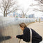A person examines a reflective memorial wall inscribed with names, with a city skyline and bare trees in the background.