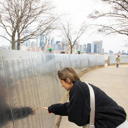A person examines a reflective memorial wall inscribed with names, with a city skyline and bare trees in the background.