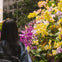 A person with long dark hair stands in front of a vibrant display of yellow and purple orchids.
