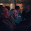 A group of people seated indoors, smiling and enjoying a performance or event, with dim lighting and a window in the background.
