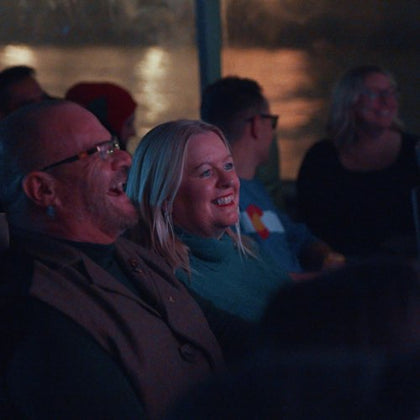 A group of people seated indoors, smiling and enjoying a performance or event, with dim lighting and a window in the background.
