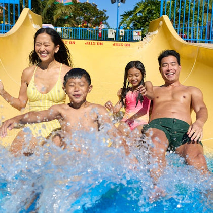 A family of four in swimwear splashing down a yellow water slide, smiling and enjoying the ride. Trees and blue railings in the background.
