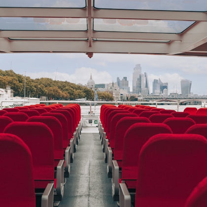 Empty rows of red seats on a boat with a glass roof, city skyline and trees visible in the background.