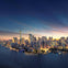 A city skyline at dusk with illuminated buildings along a waterfront, under a twilight sky.