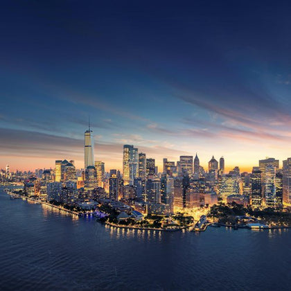 A city skyline at dusk with illuminated buildings along a waterfront, under a twilight sky.