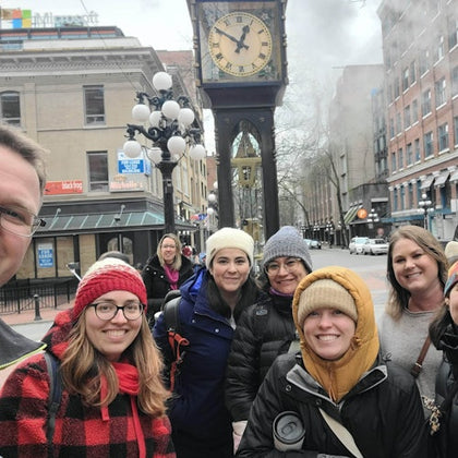 A group of people smiling in front of the Gastown Steam Clock in a city street with buildings and a lamppost visible.