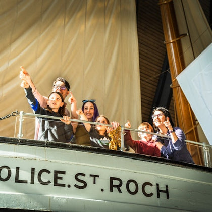 A group of people stand on the deck of a ship labeled "Police St. Roch," pointing and smiling in a bright indoor environment.