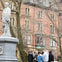 A group of people stands near a statue in a park with bare trees. Brick buildings are visible in the background.