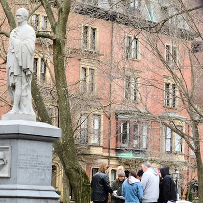 A group of people stands near a statue in a park with bare trees. Brick buildings are visible in the background.