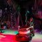 A skeleton dressed as the Grim Reaper stands in a dimly lit room with colorful lights, holding a scythe and a bumper car.