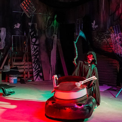 A skeleton dressed as the Grim Reaper stands in a dimly lit room with colorful lights, holding a scythe and a bumper car.