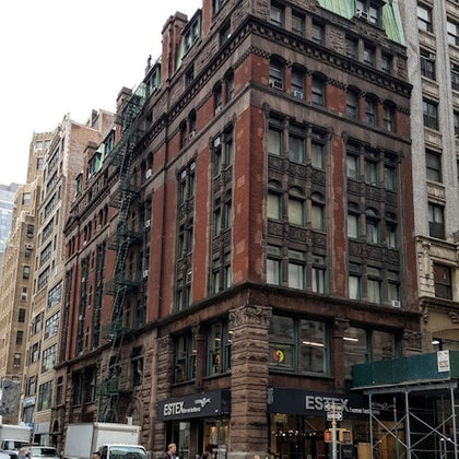 A multi-story red and brown brick building with green roof accents, featuring shops at street level, located in an urban setting.