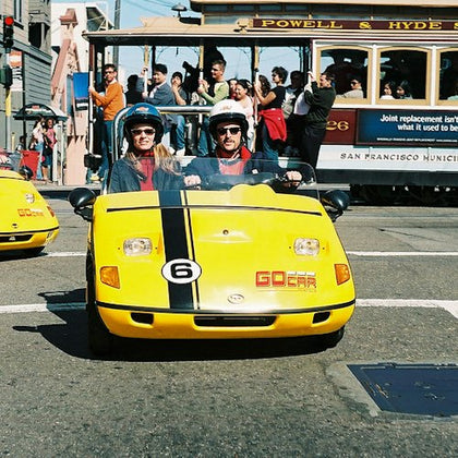 Two people in helmets are riding a yellow GoCar on a street. A Powell & Hyde cable car with passengers is in the background.