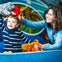 A child in a striped shirt holds a stuffed animal as an adult smiles, adjusting another plush toy on his head in a colorful setting.