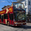 A red and yellow double-decker tour bus with passengers on the upper deck is parked on a city street under clear skies.
