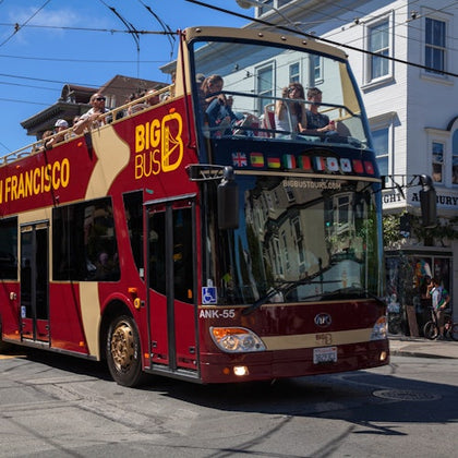 A red and yellow double-decker tour bus with passengers on the upper deck is parked on a city street under clear skies.