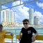 A person wearing sunglasses sitting on a boat with a cityscape of tall buildings and water in the background.