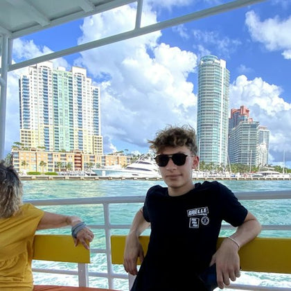 A person wearing sunglasses sitting on a boat with a cityscape of tall buildings and water in the background.