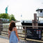 A woman stands near a dock with a view of the Statue of Liberty, a ferry named 'Miss Gateway,' and a sign for Ellis Island.