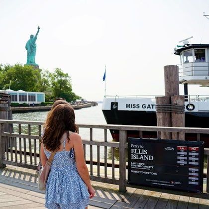 A woman stands near a dock with a view of the Statue of Liberty, a ferry named 'Miss Gateway,' and a sign for Ellis Island.