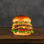A double cheeseburger with lettuce, tomato, pickles, and sauce on a wooden surface, against a dark gray background.