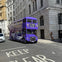 A purple double-decker bus on a city street near tall buildings. A person is standing by a wall on the sidewalk.