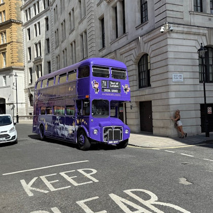 A purple double-decker bus on a city street near tall buildings. A person is standing by a wall on the sidewalk.