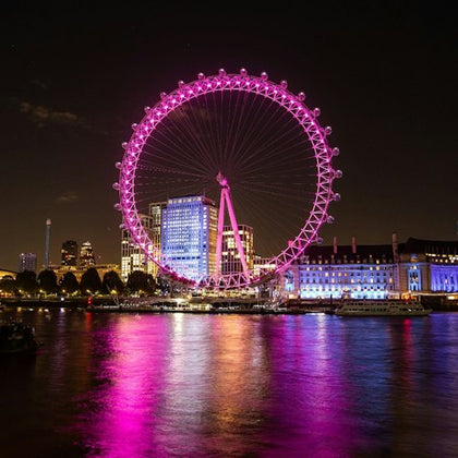 A brightly illuminated Ferris wheel with pink lights, set against a city skyline at night, reflecting on the water below.