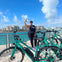 A person takes a selfie near turquoise bikes by a waterfront, with city buildings and a partly cloudy sky in the background.
