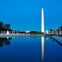 The Washington Monument reflected in a calm pool of water, with surrounding structures and a clear evening sky.