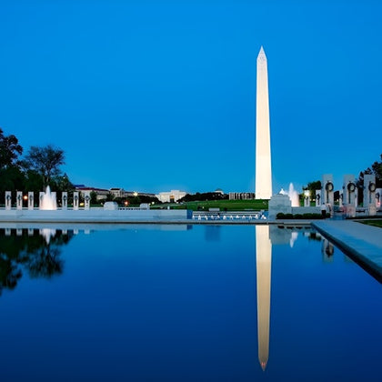 The Washington Monument reflected in a calm pool of water, with surrounding structures and a clear evening sky.
