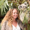 A person with curly hair smiling next to a koala on a tree surrounded by eucalyptus leaves.
