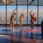 Four people stretching on yoga mats in a glass-walled room overlooking a city and water under a clear sky.