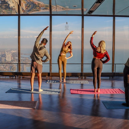 Four people stretching on yoga mats in a glass-walled room overlooking a city and water under a clear sky.