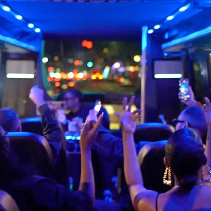 People inside a party bus at night, raising their hands and taking photos, with colorful lights and a blurred cityscape outside.