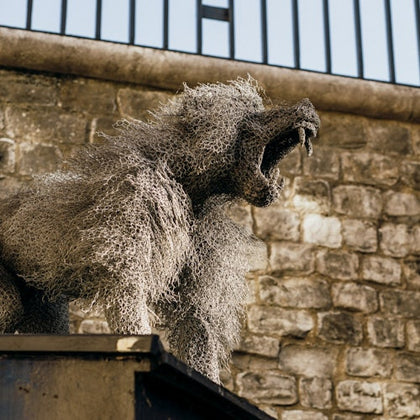 A wire mesh sculpture of a roaring lion stands against a brick wall and a metal fence.
