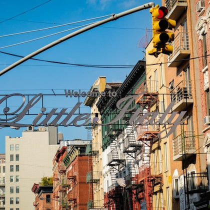 Sign reading "Welcome to Little Italy" strung between buildings, with a traffic light and fire escapes under a blue sky.