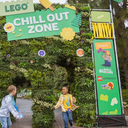 Two children happily entering a LEGO-themed 'Chill Out Zone' decorated with greenery and colorful LEGO signage.