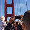 People taking photos of the Golden Gate Bridge with their phones on a sunny day. The upper part of the bridge is prominently visible.