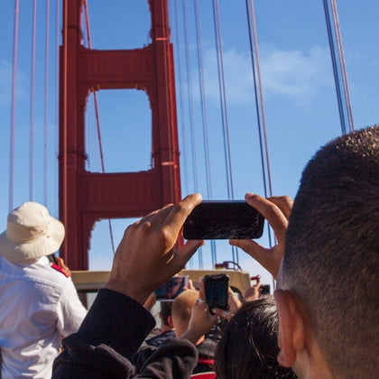 People taking photos of the Golden Gate Bridge with their phones on a sunny day. The upper part of the bridge is prominently visible.