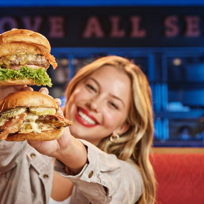A smiling person with long blonde hair holds up two large burgers, one in each hand, inside a restaurant with blue lighting.