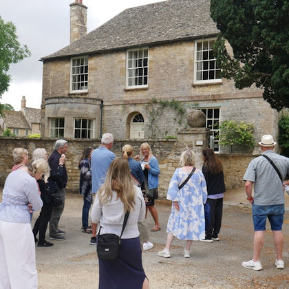 A group of people gathered outside a historic stone house with a tiled roof and large windows, surrounded by trees and greenery.