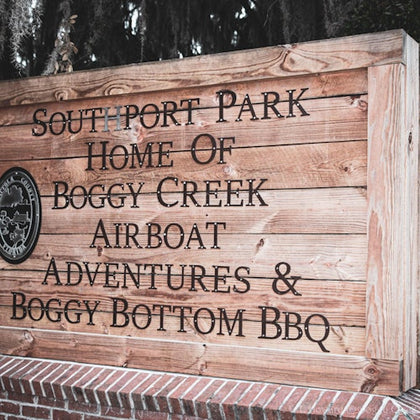 Wooden sign with text “Southport Park Home of Boggy Creek Airboat Adventures & Boggy Bottom BBQ” mounted on a brick base.