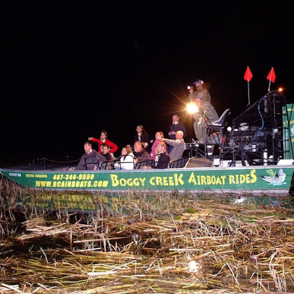 Group of people on a "Boggy Creek Airboat Rides" airboat at night, surrounded by water and reeds, with a guide standing.