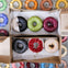 Two boxes each containing three colorful donuts, with assorted glazes, sit on a table covered in a colorful donut-patterned cloth.