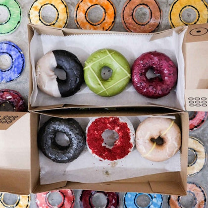 Two boxes each containing three colorful donuts, with assorted glazes, sit on a table covered in a colorful donut-patterned cloth.