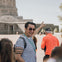 A man in sunglasses and a striped shirt gestures while leading a tour group near an ancient stone building.