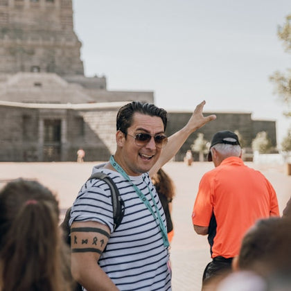 A man in sunglasses and a striped shirt gestures while leading a tour group near an ancient stone building.