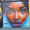 A vibrant mural on a brick wall depicting a close-up of a woman's face with expressive eyes, a nose ring, and colorful tones.