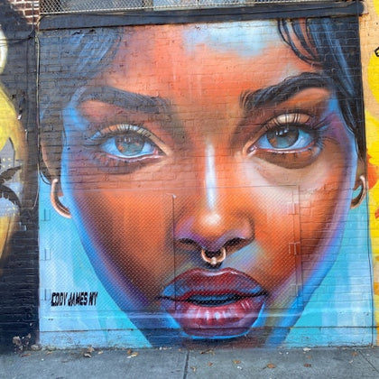 A vibrant mural on a brick wall depicting a close-up of a woman's face with expressive eyes, a nose ring, and colorful tones.